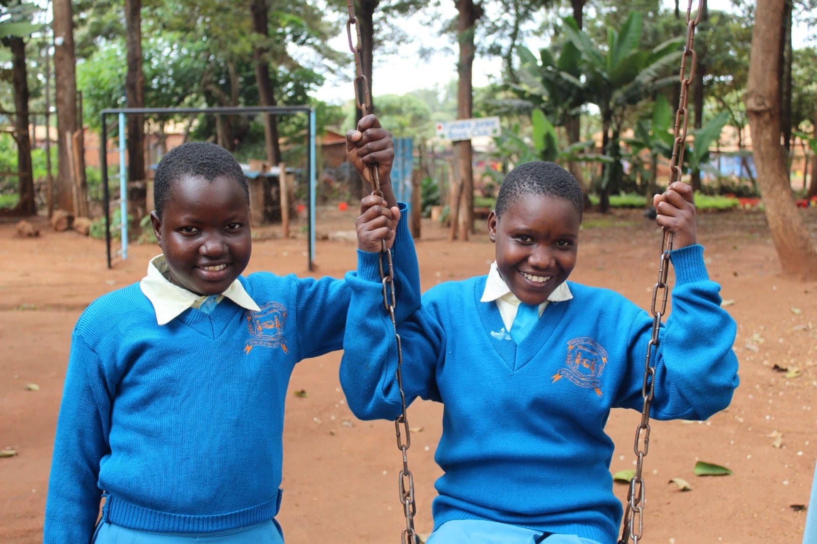 Two students smile by a swingset; Tanzania Education Corporation provides opportunities to learn digital literacy in Tanzania.