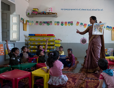 Interior scene of classroom in a government school in India. Teacher wearing a sari is reading aloud to young children sitting on the floor.